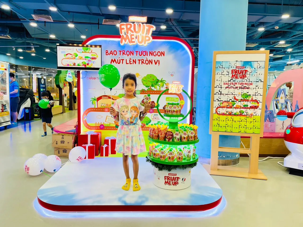 Bé đứng trên bục booth Fruit Me Up trong khu vui chơi, bên cạnh cây trưng bày sản phẩm và bảng game xếp hình