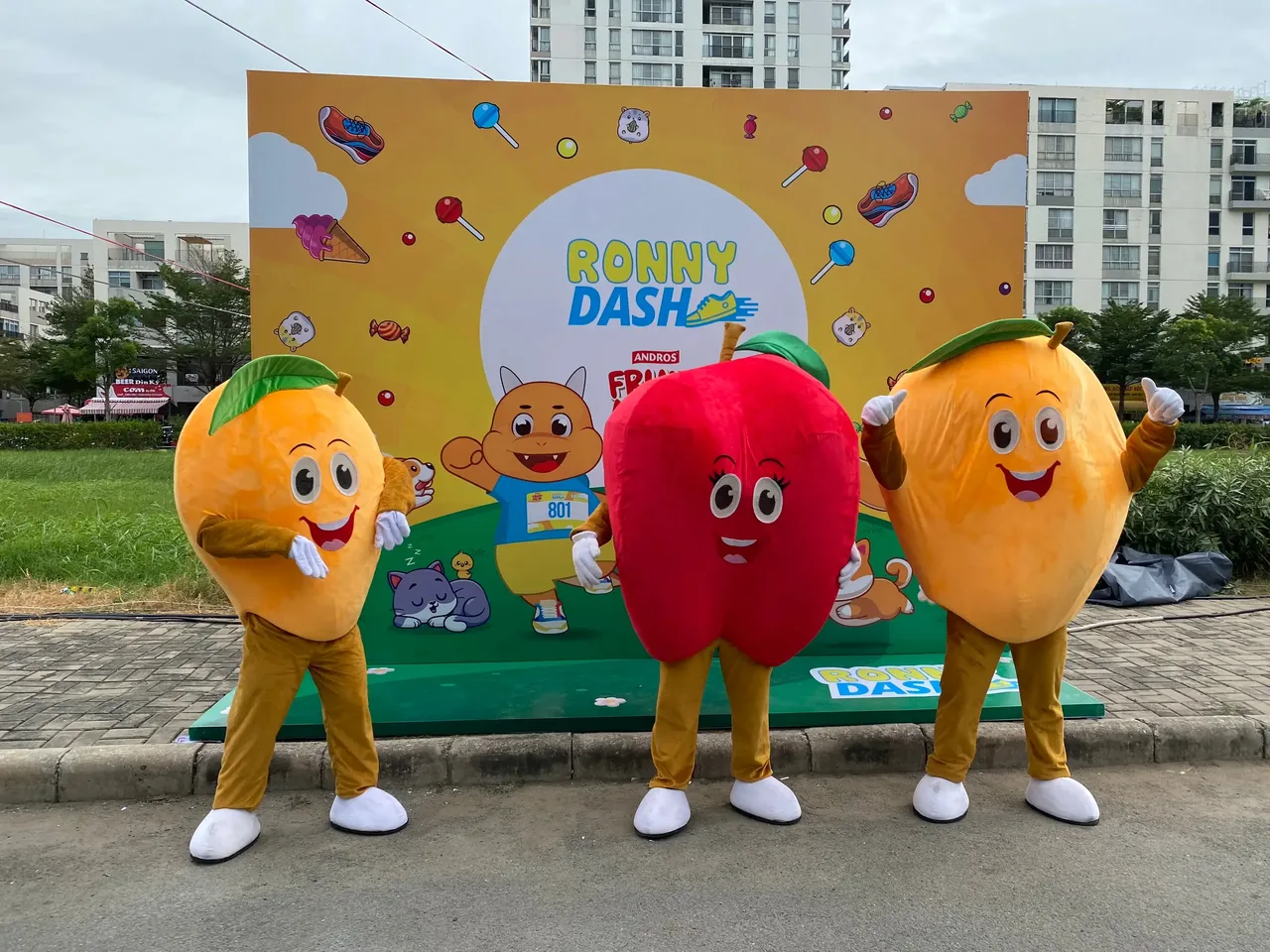 Ba mascot trái cây Fruit Me Up đứng trước backdrop Ronny Dash