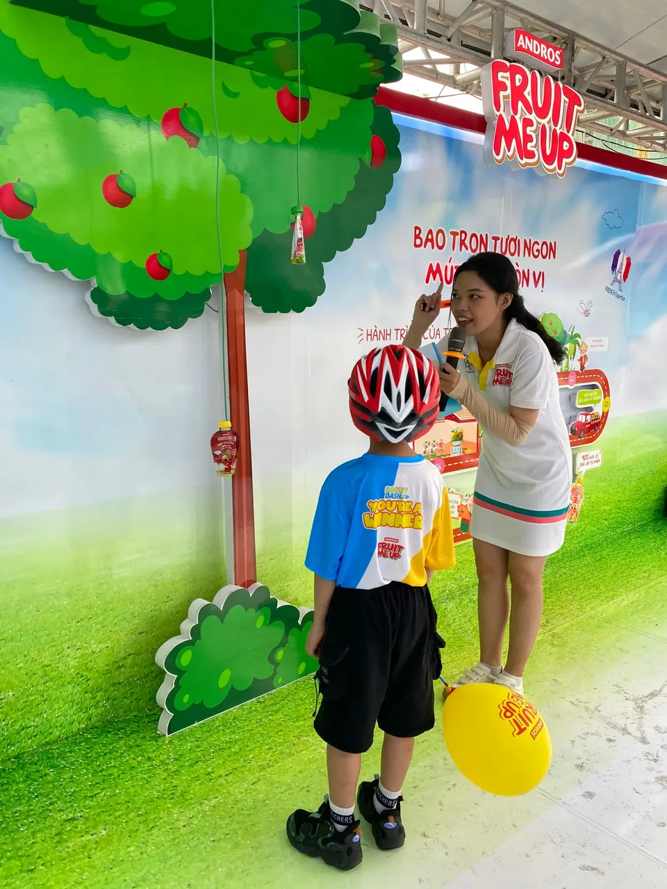 Bé đội nón bảo hiểm chơi game đánh đầu dưới cây táo của booth, PG cầm micro hướng dẫn