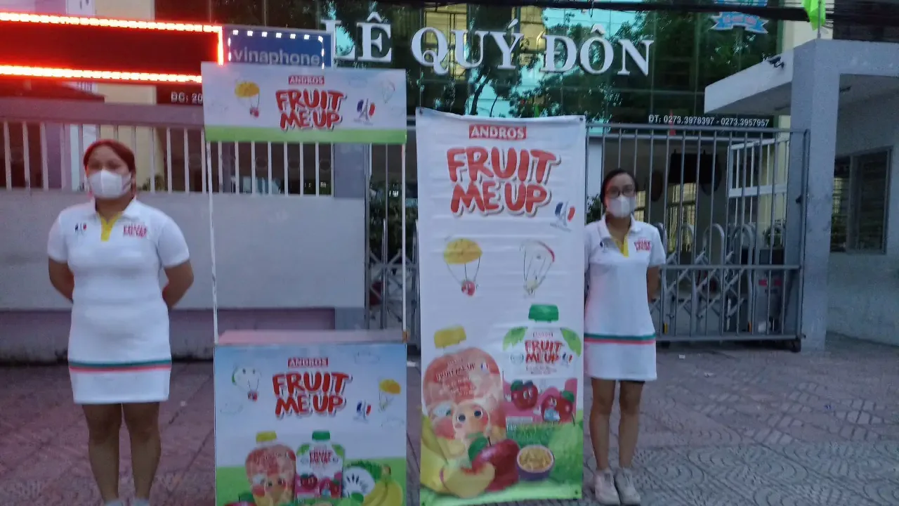 Hai PG Fruit Me Up đứng cạnh standee trước cổng trường tiểu học Lê Quý Đôn, Tiền Giang