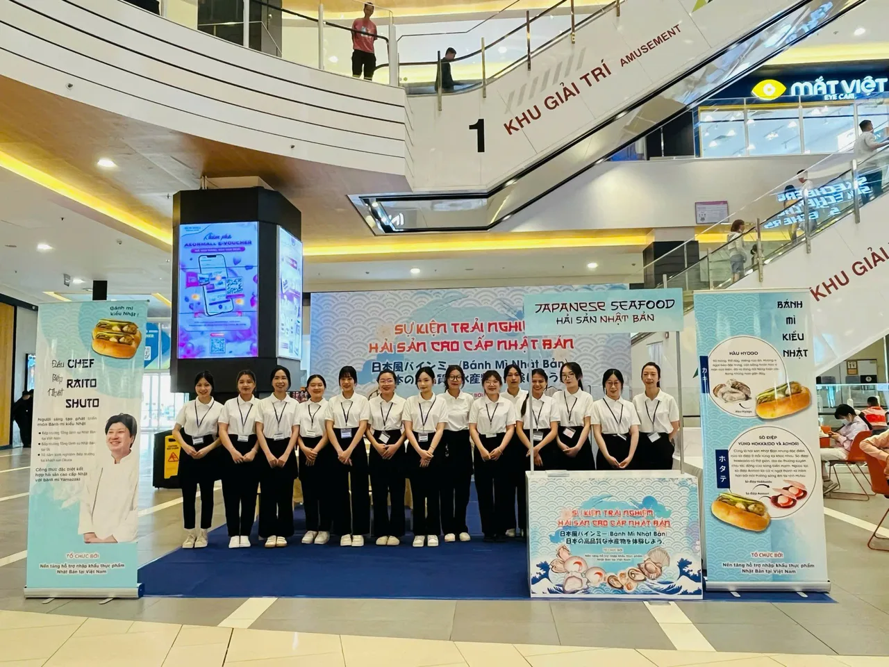 Đội PG mặc áo trắng đứng trước backdrop và các standee của chương trình tại AEON Mall Bình Tân