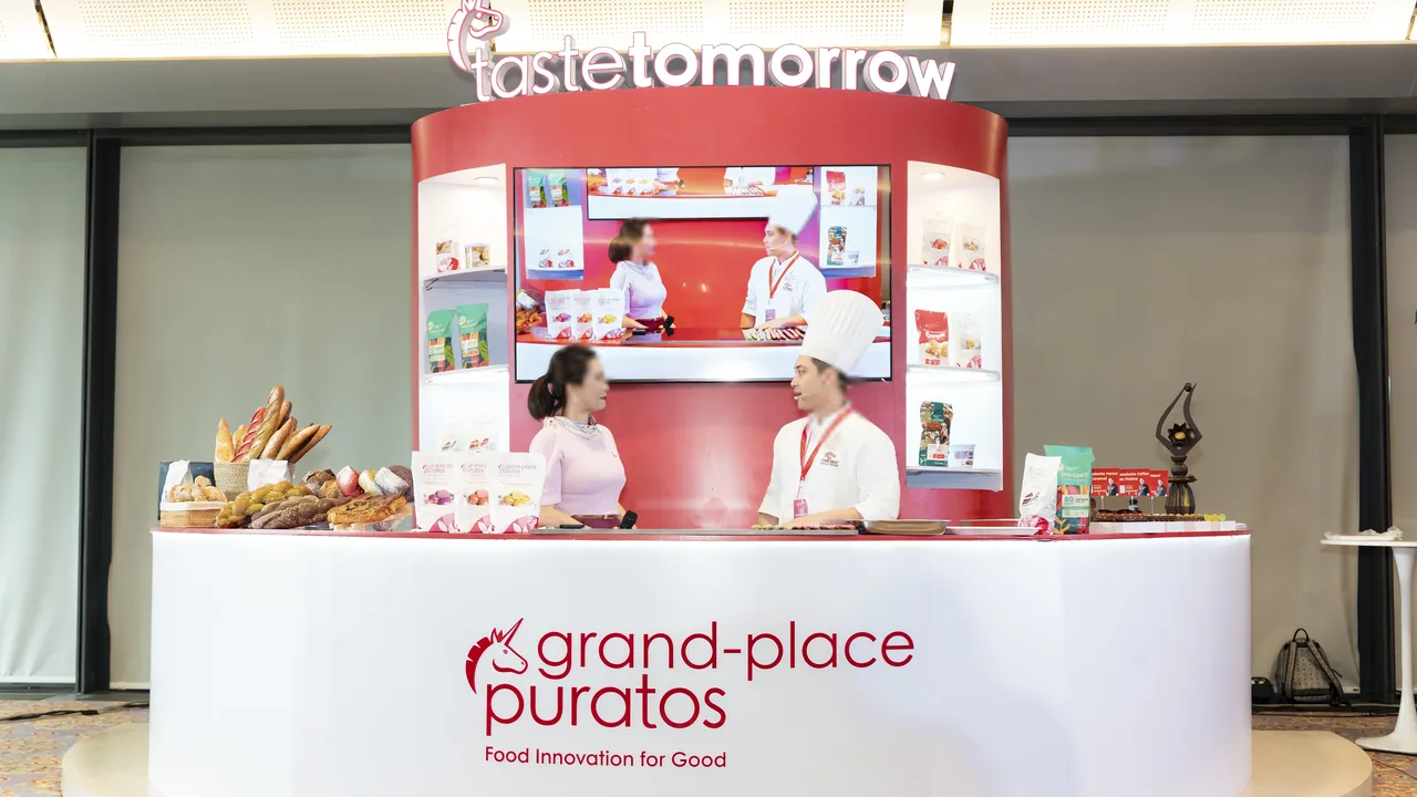 Booth trung tâm Taste Tomorrow với quầy bếp demo, màn hình lớn và kệ trưng bày sản phẩm Puratos Grand-Place