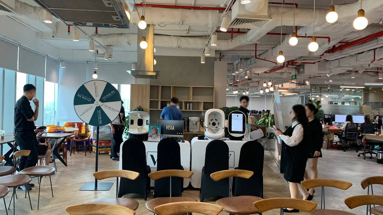 Toàn cảnh booth đợt tháng 7 với hai máy soi da, bàn branding, ghế bọc đen và vòng xoay giữa văn phòng